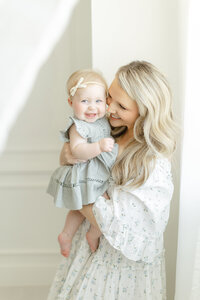 A smiling mommy and me photo taken in a DFW photography studio of a mother and her baby standing by the window as they both are smiling for their motherhood session.