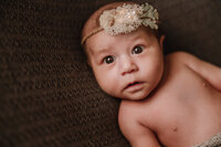 A newborn baby with a coral accented headband lies on a brown backdrop. She stares straight ahead.