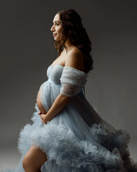 Side profile of a pregnant woman in a light blue tulle gown, highlighting her baby bump during a maternity photoshoot by Kaitlyn Dawn Photography in the Inland Empire.