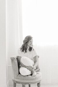 Black and white in studio professional headshot photo of a woman sitting down on a chair while she looks over her shoulder smiling.
