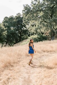 Brunette girl wearing a royal blue dress and looking over her shoulder while walking through tall, dry grass