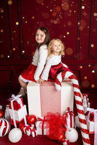 Two sisters sit on a large white box wrapped in red ribbon. They smile cheerfully. They're surrounded by red and white gift boxes and ornaments.