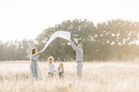 A family photo of mom an dad holding a blanket up in the air as their boys run under in the middle of a Dallas/Frisco TX field.