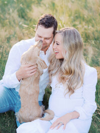 Pregnant woman, her husband and their dog are sitting on the ground and laughing as the dog licks husband's nose