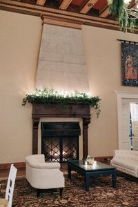 Ornate living room with a large vintage fireplace, decorated with greenery, flanked by elegant furniture and an ornamental rug.