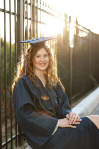 High school graduate in a cap and gown sitting by a wrought iron fence with warm sunset light, senior graduation portrait by Kaitlyn Dawn Photography.