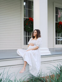 Pregnant woman sitting on a white porch holding her pregnant body