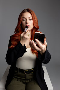 Woman with long red hair smiling while holding mascara and compact mirror. Photograph by Yucaipa branding photographer Kaitlyn Dawn Photography.