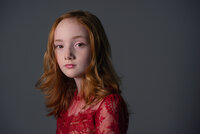 Portrait of a young girl with long red hair in a red lace dress against a dark grey backdrop.