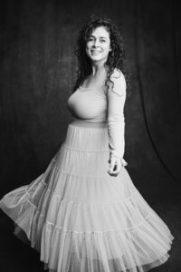 A black and white picture of a woman with dark curly hair smiling widely as she twirls in front of a dark gray backdrop. She wears a long tulle skirt that fills the lower half of the frame.