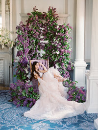 bride seated in a velvet chair with flowing gown and large lilac installation in a French-inspired interior
