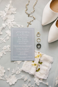 Flat lay of light blue wedding stationery surrounded by wedding rings, a handkerchief, and white heels