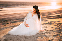 A pregnant woman with dark hair sits on the back staring down at her baby bump. She cradles her bump with one hand while the other hand rests behind her, helping her support herself. The sun is setting over the beach behind her.