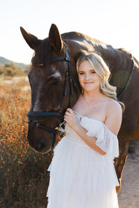 High school senior girl in a white dress standing beside a horse during golden hour, outdoor senior portrait by Kaitlyn Dawn Photography.