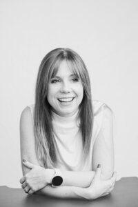 A black and white picture of a blonde woman with straight hair sitting in front of a white backdrop. She wears a white shirt and smiles brightly. Her arms are crossed on the black table in front of her.