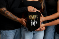 Close-up of a pregnant woman’s belly with family hands holding a “Native Baby” outfit, capturing a meaningful pregnancy moment during a Yucaipa session by Kaitlyn Dawn Photography.