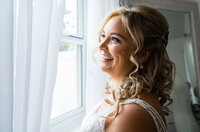 Bride smiling by a window with soft natural light highlighting her curled hair and lace wedding dress during her getting-ready photos.