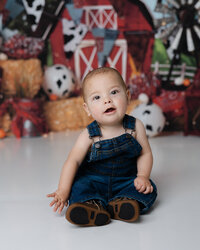 Baby boy sitting in front of a farm-themed backdrop wearing denim overalls during his first birthday photo session.
Photograph by Yucaipa cake smash photographer Kaitlyn Dawn Photography.