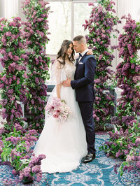 bride and groom standing in a dramatic lilac floral aisle with layered blooms and soft window light behind