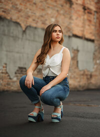 Senior portrait in downtown Mobile, Alabama — crouching in front of a large weathered brick wall looking off in the distance.