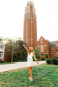 Girl in white dress holding graduation cap standing on lawn outside of Vanderbilt tower