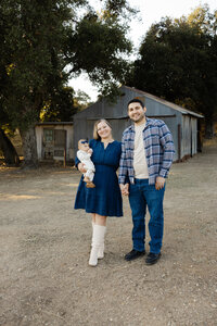 Young family of three standing outdoors in front of a rustic barn, neutral coordinated outfits and natural light captured by Kaitlyn Dawn Photography, professional outdoor family portrait photographer.