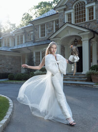 Model in a white gown with a sheer overlay moving through the forecourt of a stone manor house.