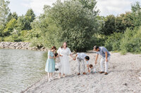 A family gathers on the rocky lakeside shore at Colonel Samuel Smith Park, with trees and bushes framing the scene. Grandparents enjoy moments interacting with small children, creating a perfect snapshot of joy and togetherness.
