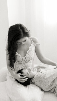 Black and white portrait of a mother sitting on the edge of a bed as her son lays on her lap while she holds him.