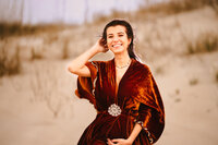 A pregnant woman in an orange velvet dress smiles against a sandy beach dune.