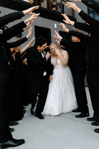 Bride and groom laugh and celebrate walking through cheering wedding party in black formal attire.