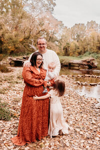 Mom, dad, and two children smiling on a creekside.  mom is wearing burnt orange dress from Baltic Born, and daughter is wearing ivory dress from Joyfolie.  Photo taken by philadelphia family photographer, Kristi
