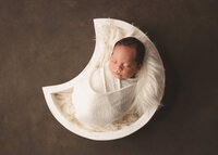 Moon shaped newborn prop with baby boy on brown backdrop.
