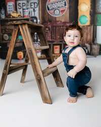 Happy one-year-old boy posing with a small wooden ladder in front of a vintage truck-themed cake smash set. Photograph by Yucaipa cake smash photographer Kaitlyn Dawn Photography.
