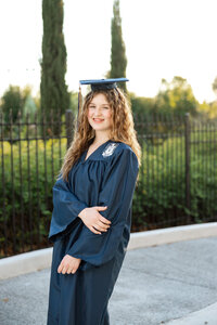 High school graduate wearing a blue cap and gown standing confidently outdoors near a fence and trees, senior portrait by Kaitlyn Dawn Photography.