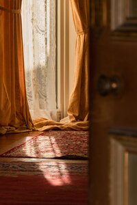 Long, floor length tan curtains hand parted before a glowing window casting a lightbeam across a warm, red rug.
