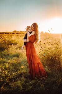 a woman in a red dress standing in a field holding her child in a close hug with the sun setting behind them during their family photoshoot.