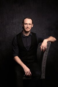 A dark haired man with light blue eyes smiles softly. He sits sideways in a gray linen chair with one arm draping the the chair back and the other laying across his lap. He wears a gray shirt with a black vest and sits in front of a gray backdrop. The lighting is dramatic.