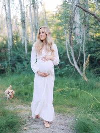 Expectant mom is walking in the woods with her dog, while holding her bump