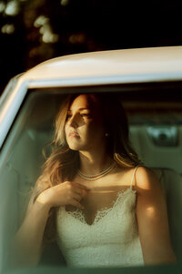 Bride in lace gown sitting in vintage car looking outside with golden sunlight on wedding day.