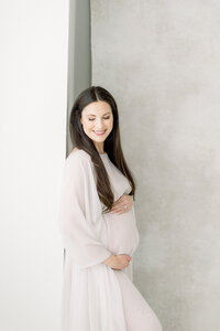Photo of an expecting Dallas mom leaning on a wall while she is dressed in a tulle maternity gown holding her belly in a Dallas photography studio.