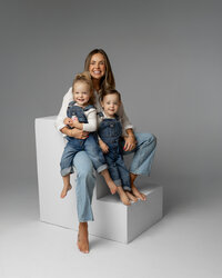 Mother sitting on white studio blocks cuddling her two toddlers, soft neutral tones and modern studio styling by Kaitlyn Dawn Photography, professional family photographer.