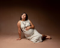 Pregnant mom reclining on the floor in a beige dress, softly cradling her belly during a maternity photoshoot in the Inland Empire by Kaitlyn Dawn Photography.