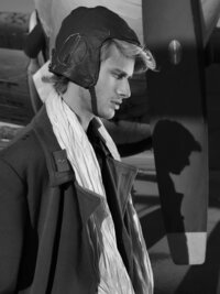Black and white profile of a young man in a vintage pilot cap and coat, his shadow cast on the wall behind.