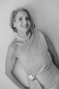 A smiling woman with short hair wears a one-shoulder textured gown, multi-strand pearl necklace, and large hoop earrings. Posing against a plain background, she embodies the elegance of editorial portrait photography in the Washington DC metro area.