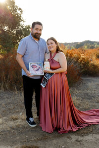 Expecting couple standing outdoors during a golden hour lifestyle maternity session holding ultrasound photos and a framed newborn picture while celebrating their pregnancy photographed by Kaitlyn Dawn Photography.