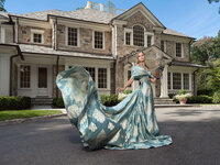 Woman in a blue-green gown with a long train standing before the entrance of a large stone manor house.