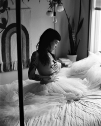 a black and white photo of a non-binary person seductively sitting in bed with no shirt on