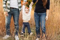 A child in a white sweater and blue pants is joyfully swinging between two adults holding hands in a grassy field, captured beautifully by a Toronto lifestyle family photographer.