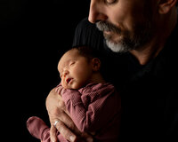 Father gently holding his sleeping newborn baby during a studio newborn photography session with soft dramatic lighting by Inland Empire newborn photographer Kaitlyn Dawn Photography.